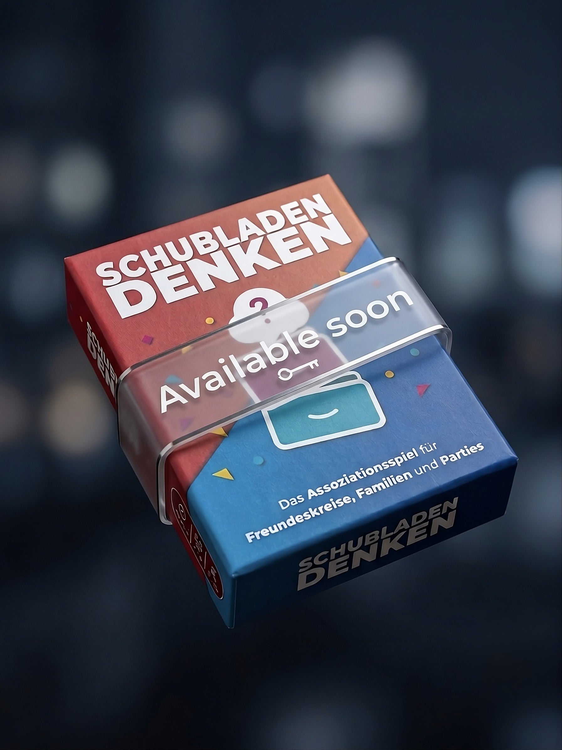 A 3D render of a product box for 'Schubladen Denken' featuring a red top half and a blue bottom half, with a clear plastic wrap displaying 'Available soon' text, set against a dark blurred background.